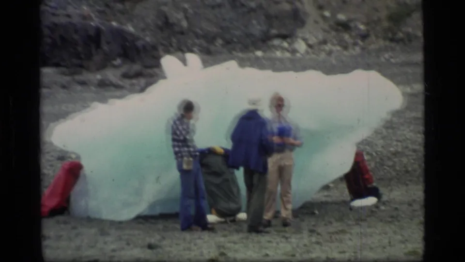 In Glacier Bay, Alaska, three adults in cold-weather clothing examine a large translucent ice block on a rocky beach.