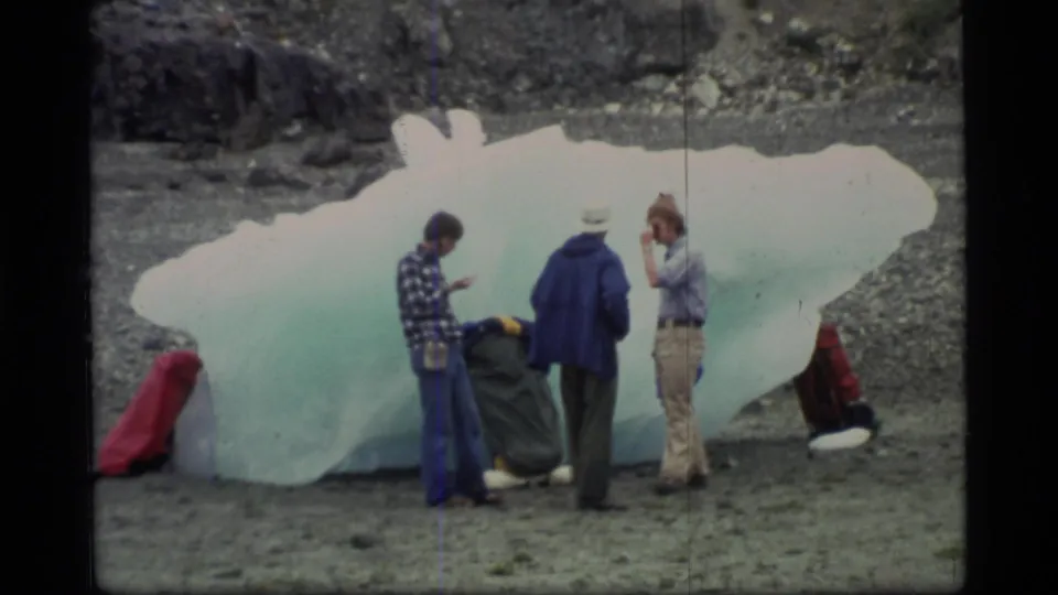 Three adults appear to inspect a blue-white ice block on the Glacier Bay shoreline, with red bags set near the ice.