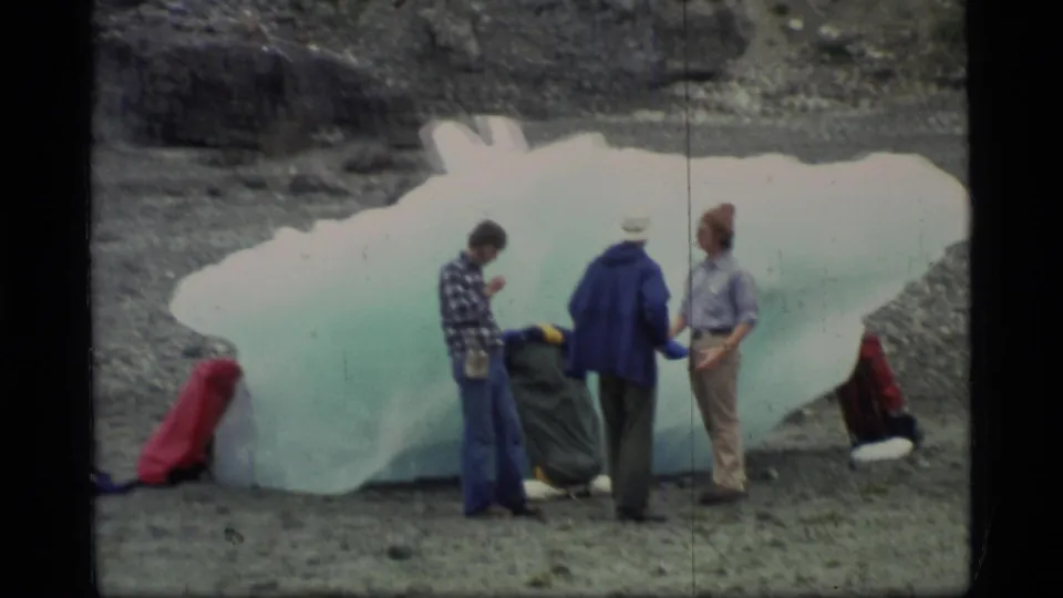 Adults on a rocky Glacier Bay beach stand close to a large pale-blue ice block, with travel bags placed beside it.