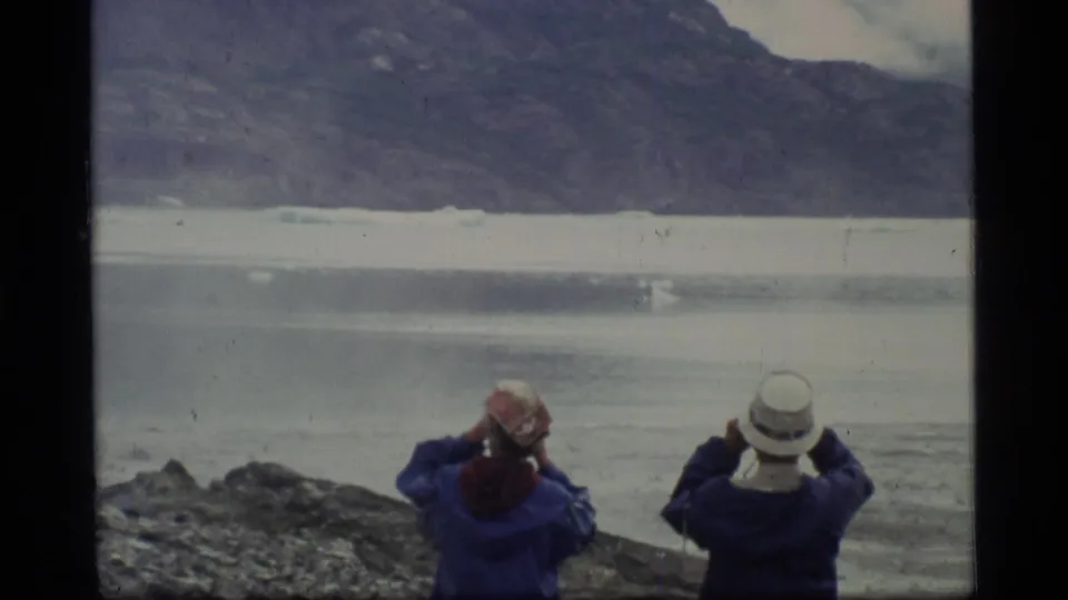Two adults in hats look out over calm Glacier Bay water toward floating ice and distant mountains.