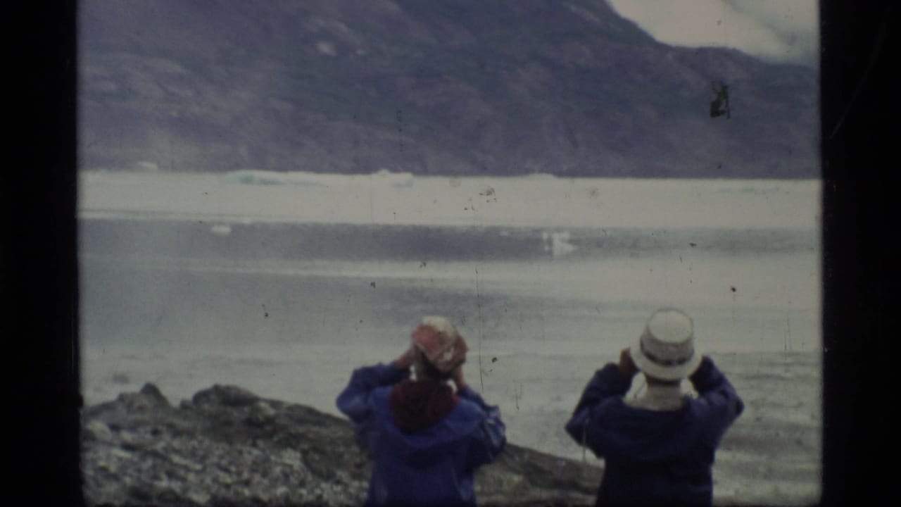 In Glacier Bay, Alaska, two travelers in blue jackets and hats face a glacial lake from a rocky shoreline in 1977 home-movie footage.