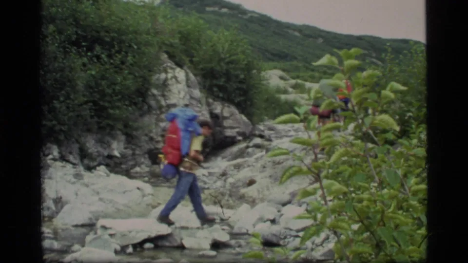 A hiker carrying a red and blue backpack crosses rocks beside a shallow stream in Glacier Bay, Alaska, in 1977 8mm footage.