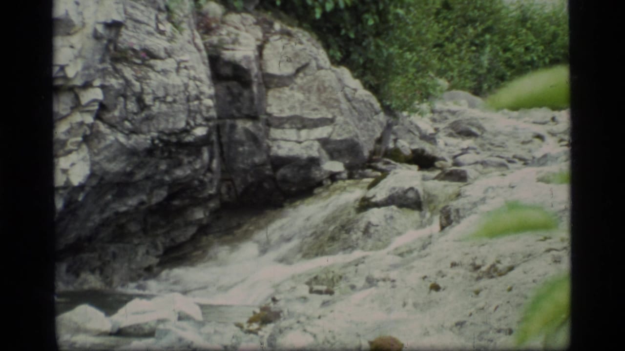 1977 8mm Glacier Bay travel footage frame showing a rocky stream, pale boulders, cliff face, and nearby green vegetation.