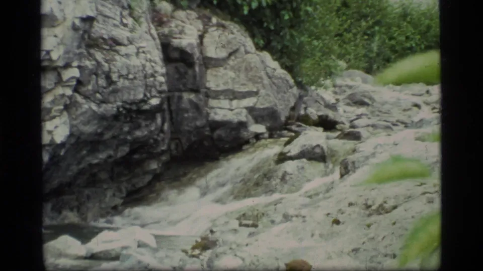 1977 8mm Glacier Bay travel footage frame showing a rocky stream, pale boulders, cliff face, and nearby green vegetation.
