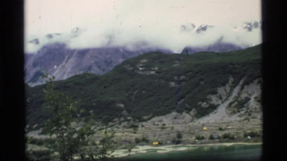1977 Glacier Bay 8mm frame looking across a lake edge toward green hills, pale slopes, and mountains partly covered by cloud.