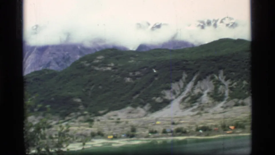 1977 8mm Glacier Bay view across water and valley toward green hills, exposed pale slopes, distant buildings, and clouded mountains.