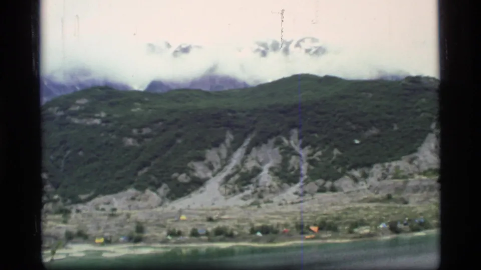 Final 1977 Glacier Bay frame showing green valley slopes, pale eroded faces, lake water, low clouds, and a small distant settlement.