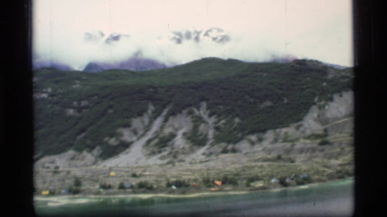 1977 Glacier Bay, Alaska home-movie frame showing a calm lake below forested slopes, pale rock scars, and clouded snow peaks.