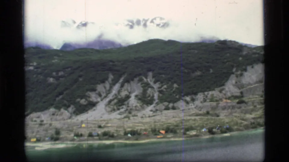 1977 Glacier Bay, Alaska frame with a mountain lake, green slopes, pale slide areas, and small colorful shoreline tents or structures.