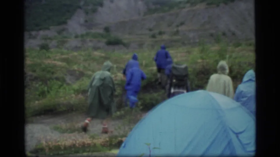 1977 Glacier Bay camping scene with several people in rain ponchos moving away from a blue tent through damp vegetation.