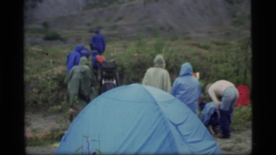 1977 Glacier Bay frame showing a blue tent, campers in ponchos, and one person bending near bags at a damp campsite.