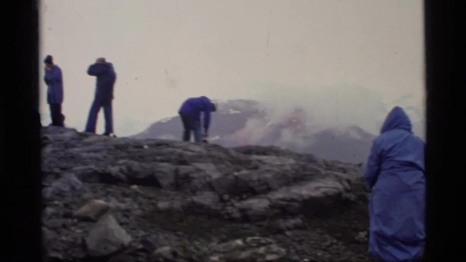 Visitors in blue raincoats appear on a rocky Glacier Bay overlook, with haze, snow patches, and distant mountains in the background.