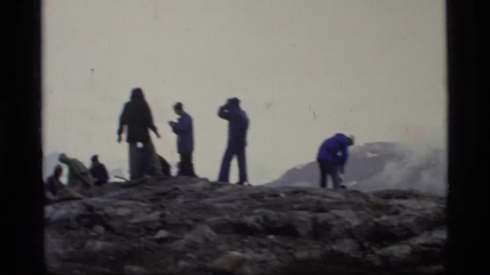 A small group of Glacier Bay visitors stands and crouches on dark rock, with blue outerwear and a hazy mountain backdrop visible.