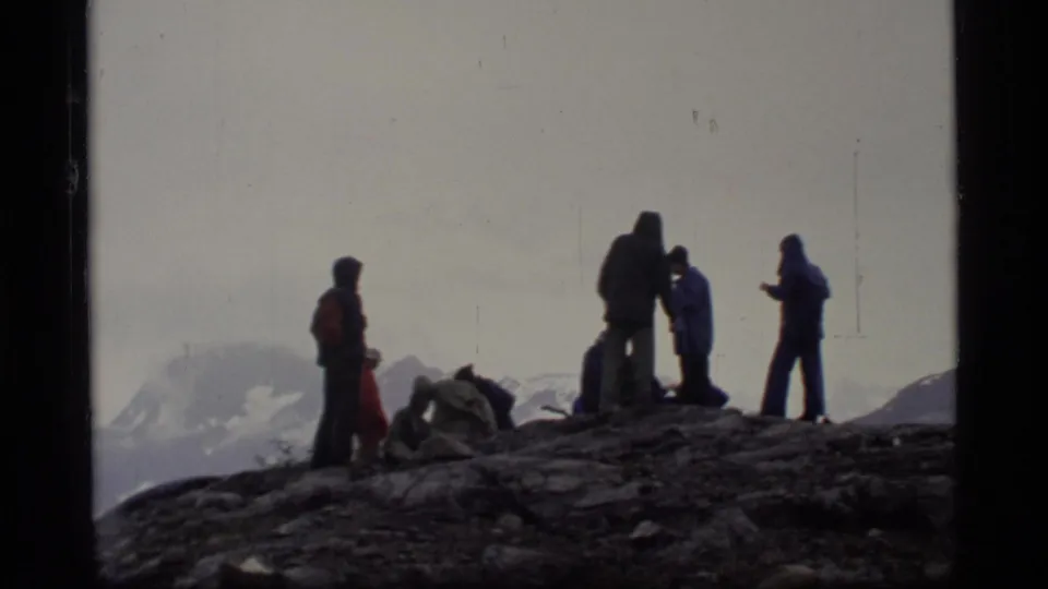 Glacier Bay visitors stand in silhouette on a rocky ridge, with mist, distant snow-covered mountains, and hooded rainwear visible.