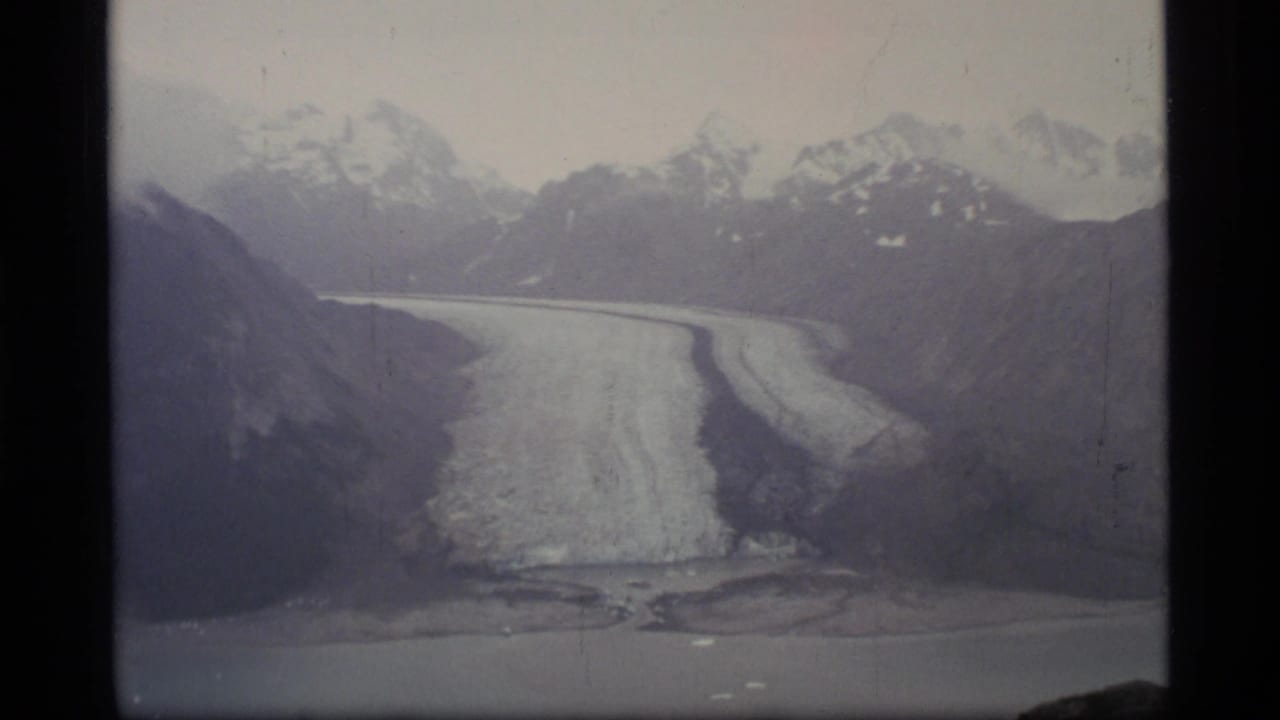 1977 8mm view of a glacier in Glacier Bay, Alaska, with snow-capped mountains behind and water in the foreground.