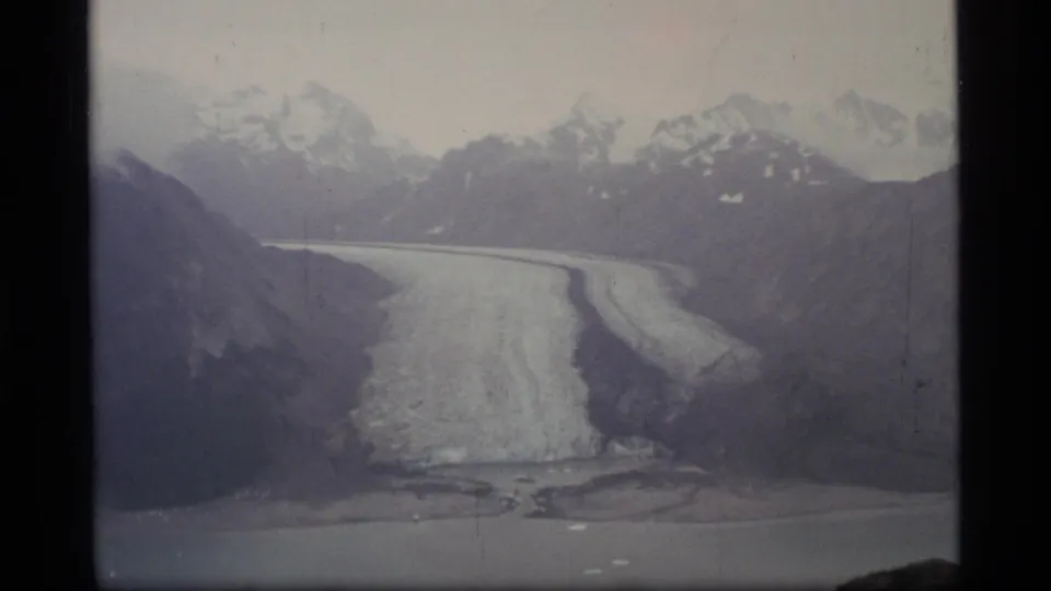 1977 8mm view of a glacier in Glacier Bay, Alaska, with snow-capped mountains behind and water in the foreground.