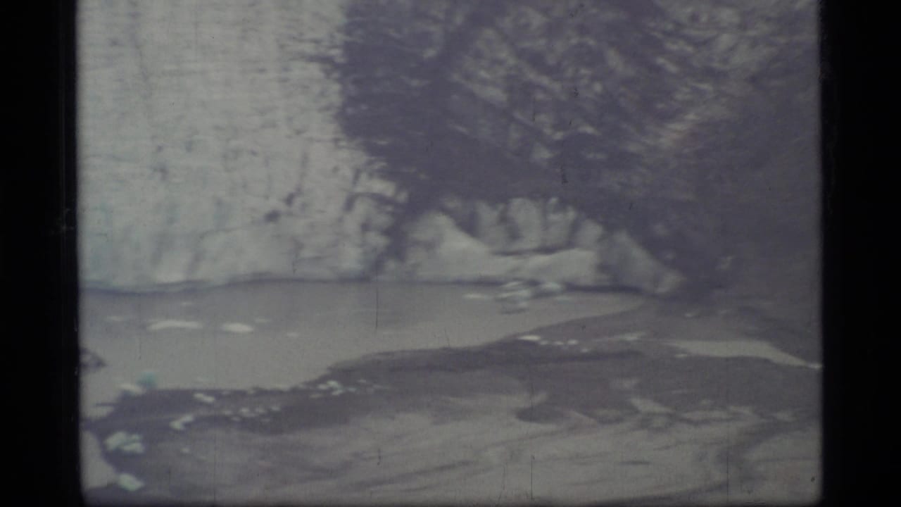 1977 8mm Glacier Bay view with icy shoreline, pale water, and dark vertical film-frame edges.
