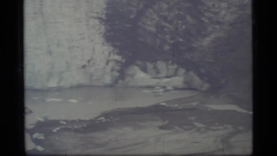1977 8mm Glacier Bay view with icy shoreline, pale water, and dark vertical film-frame edges.
