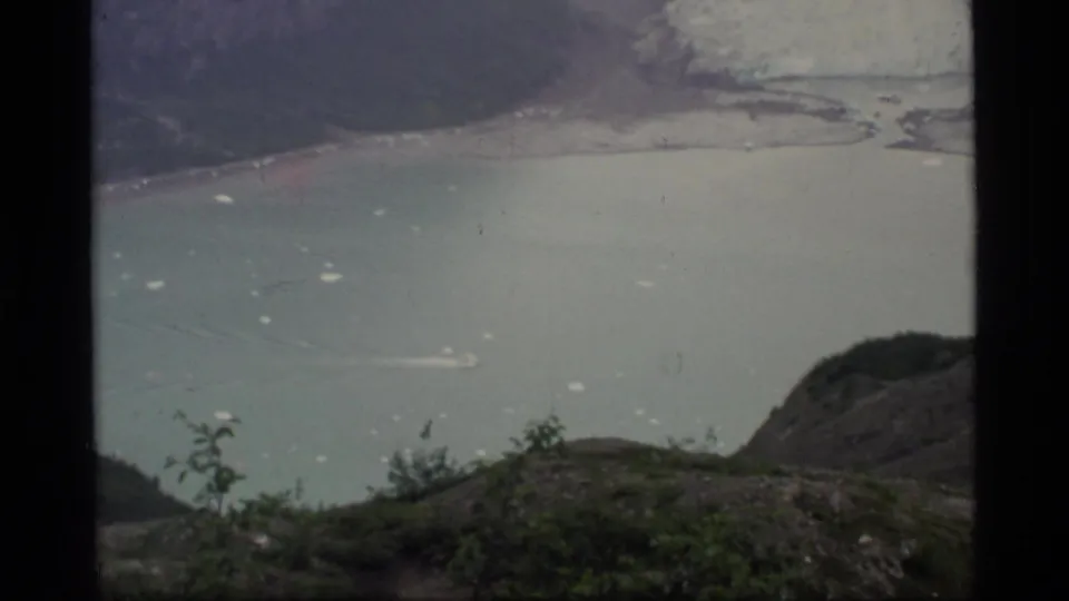 Small boat leaves a wake across Glacier Bay water below foreground brush and dark mountain slopes in 1977 home-movie footage.
