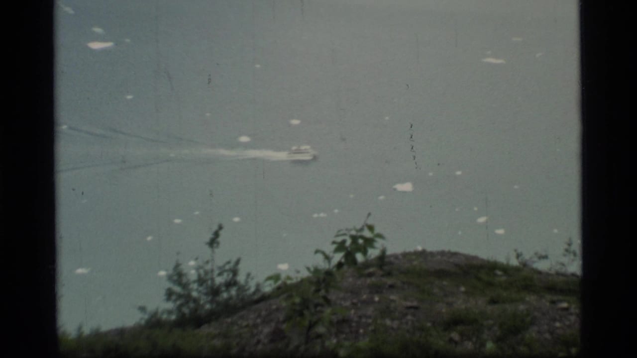 1977 8mm Glacier Bay view of a small boat moving across calm gray-blue water beyond a rocky, shrub-covered foreground.
