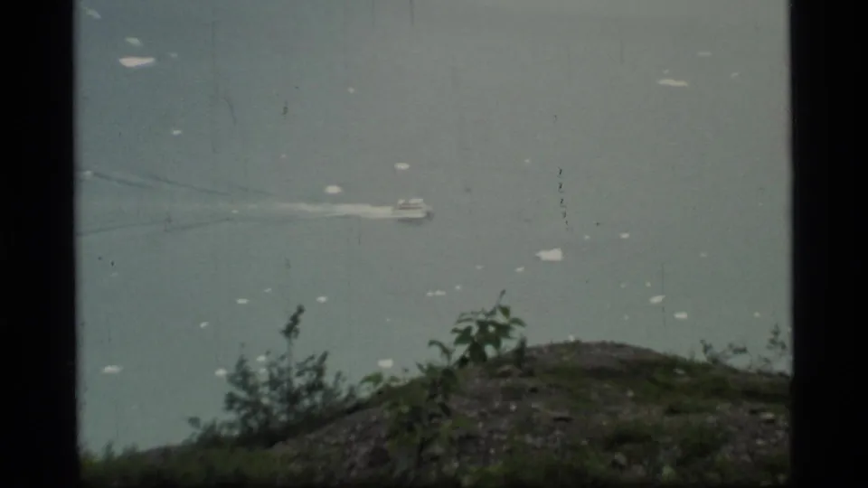 1977 8mm Glacier Bay view of a small boat moving across calm gray-blue water beyond a rocky, shrub-covered foreground.