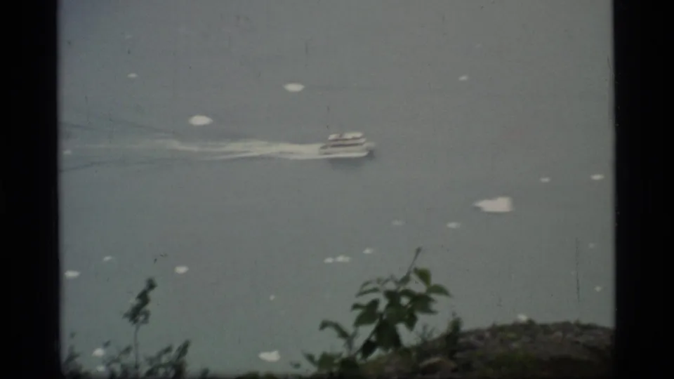Small boat and white wake on calm Glacier Bay water, seen from a high 1977 home-movie shoreline viewpoint.