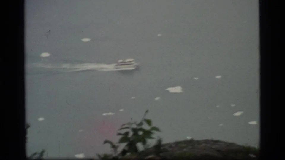 Vintage 8mm frame showing a small boat in Glacier Bay water, with scattered pale patches and a rocky green foreground.