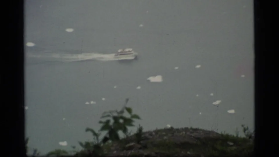 Glacier Bay home-movie frame of a small boat crossing open water, with a shrub and rocky slope in the foreground.