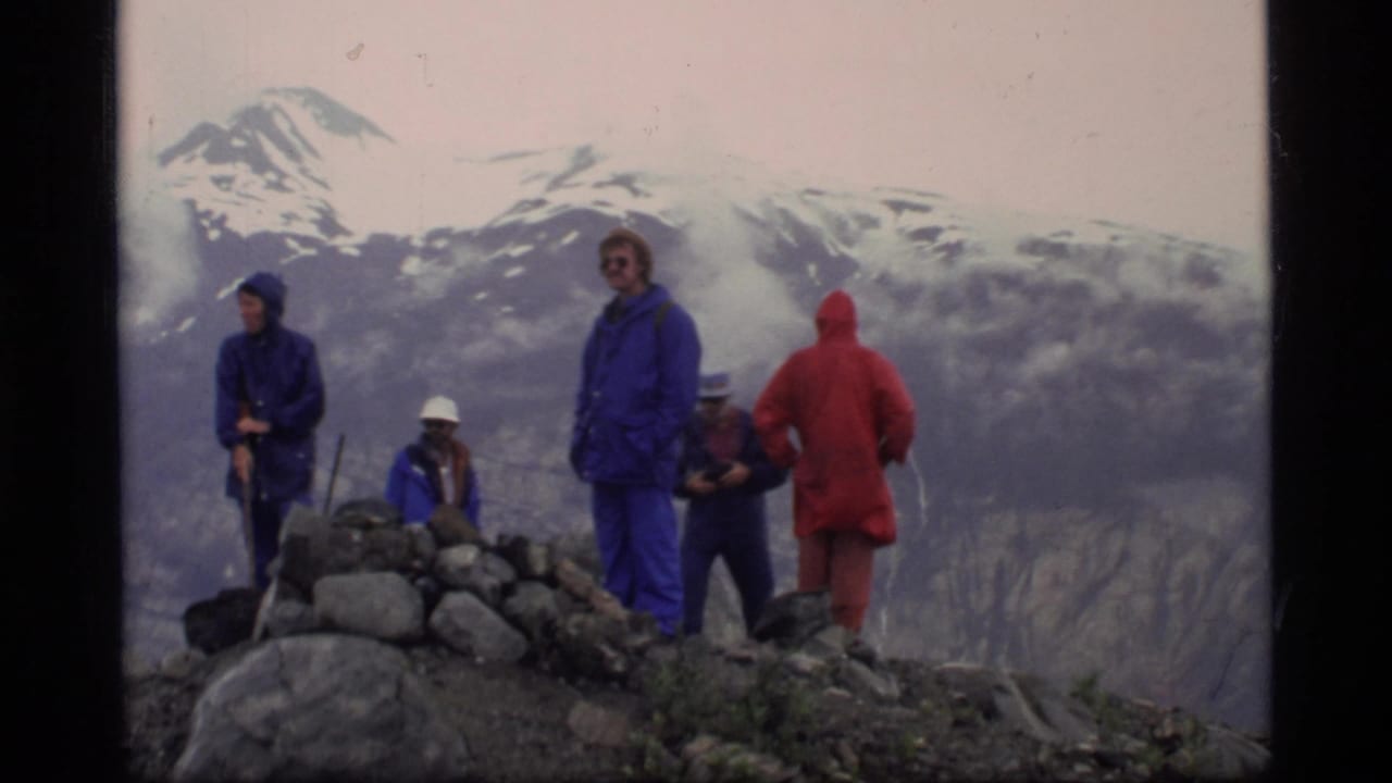 Hikers in red and blue rain gear stand on a rocky ridge with snow-capped mountains behind them.