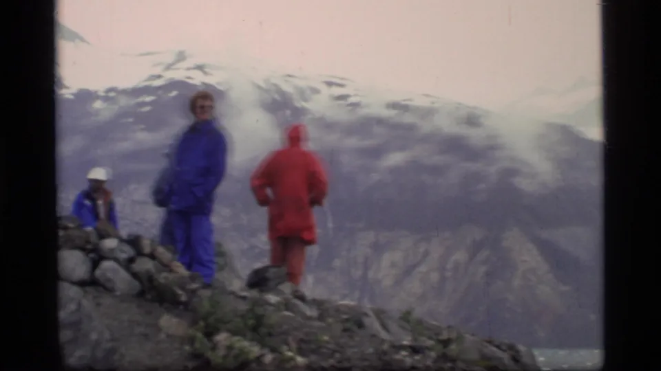 1977 Glacier Bay frame with hikers in blue and red rain gear on a ridge above distant snow-covered mountains.