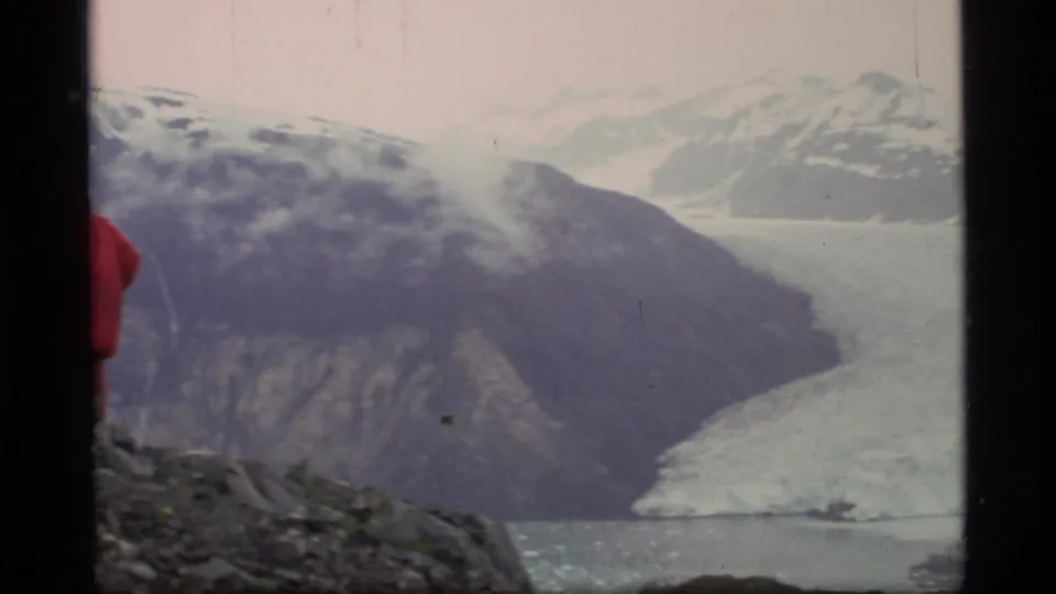 Glacier Bay 1977 home-movie frame with a broad glacier, steep mountain slopes, water, and a partial red parka at left.