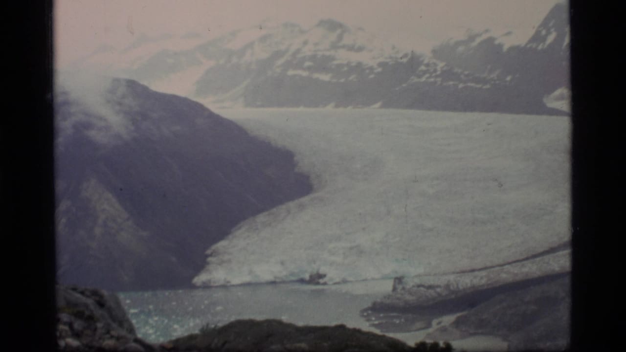 1977 8mm view of Glacier Bay, Alaska, showing a broad glacier, dark rocky slopes, snowy mountains, and cold bay water.