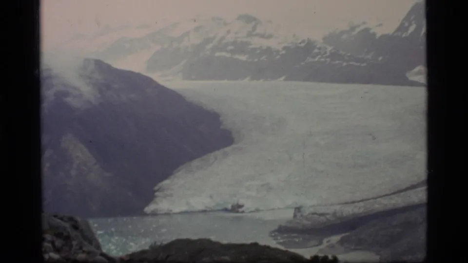 1977 8mm view of Glacier Bay, Alaska, showing a broad glacier, dark rocky slopes, snowy mountains, and cold bay water.