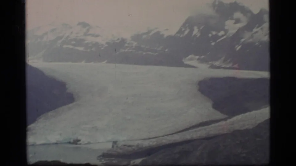 Archival 8mm frame from Glacier Bay showing a pale glacier, distant snowy ridges, dark rocky land, and cold water below.