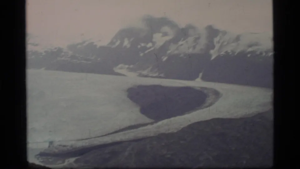 1977 Glacier Bay home-movie frame with a sweeping glacier, snowy mountain backdrop, dark rock formation, and cold water near the ice.