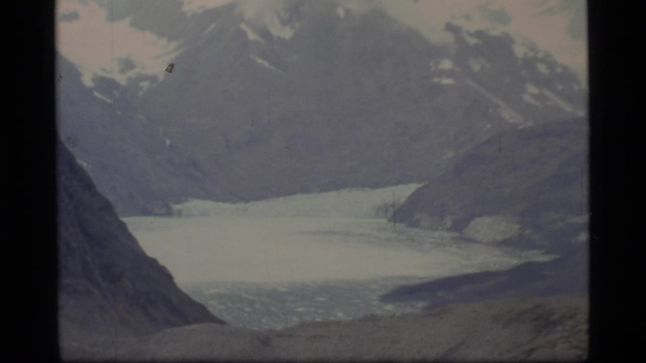 Distant glacier or frozen bay below snow-capped mountains, framed by dark rocky slopes.