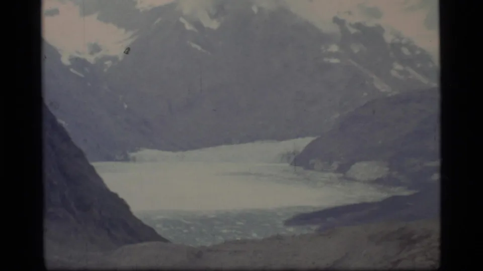 1977 Glacier Bay, Alaska 8mm frame showing a pale glacial bay beneath snow-capped peaks and dark mountain walls.