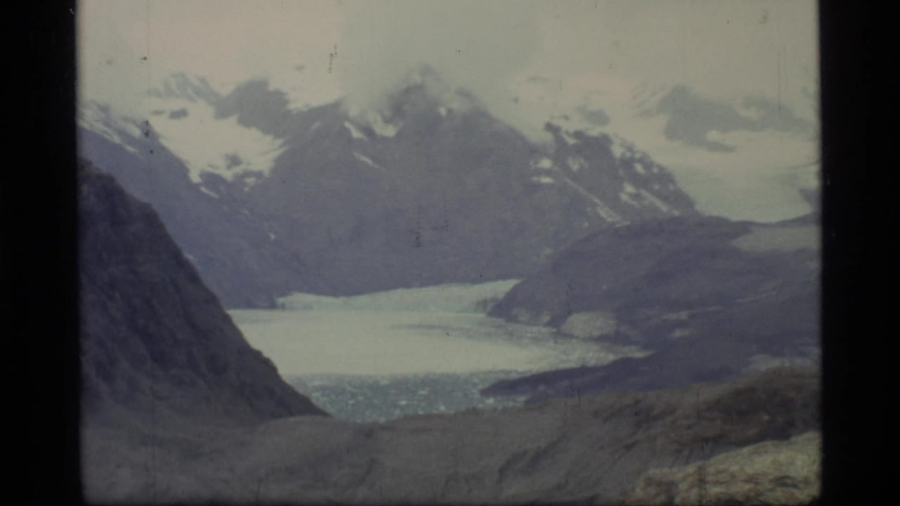 Snowy mountain range and pale glacial water seen between dark valley slopes.