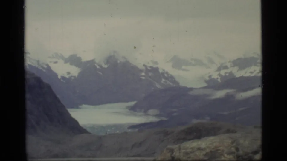 1977 home-movie view of Glacier Bay with a broad icy bay, snow-capped mountains, dark rock slope, and muted archival film color.