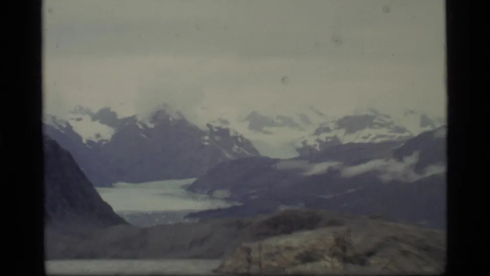 Glacier Bay, Alaska 8mm frame showing layered snow-capped mountains, pale glacier or bay ice, and rocky foreground terrain.