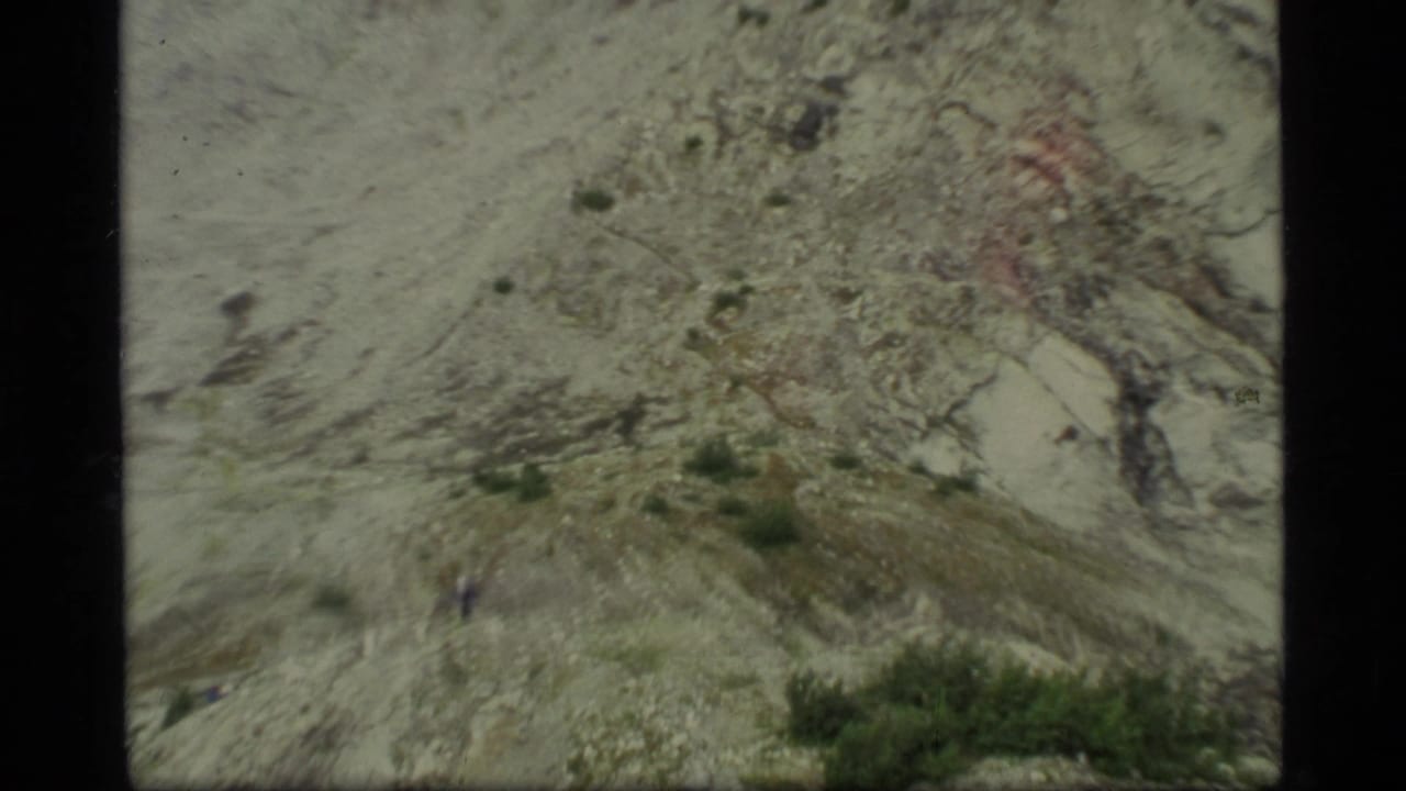 1977 Glacier Bay frame of pale rocky terrain with scattered vegetation and a small distant hiker on the slope.