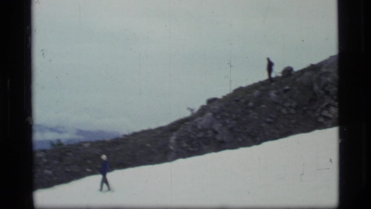 Small hikers crossing a snowy and rocky mountain slope beneath an overcast sky.