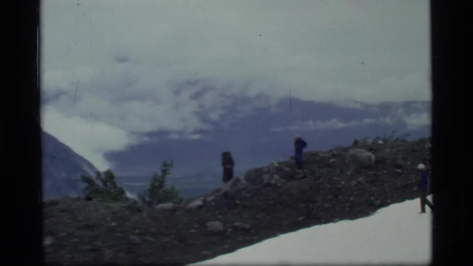 Glacier Bay hiking party seen at a distance on rocky alpine terrain, framed by snow, clouds, and muted 8mm color.