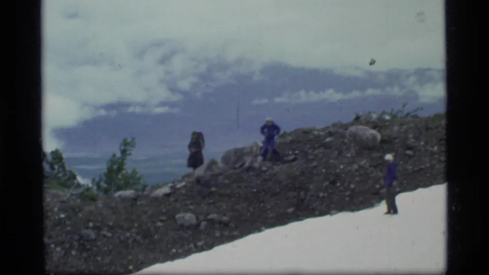 A small group of hikers in winter clothing stands on a rocky Glacier Bay slope with snow and cloudy mountain views.