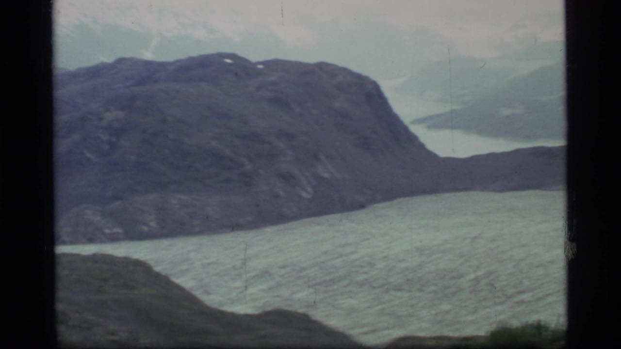 Rocky mountain above pale fjord water with distant snow and clouds.