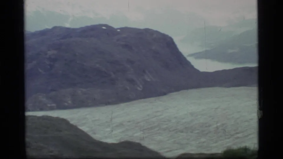 Glacier Bay, Alaska landscape view with a dark rocky mountain, pale fjord water, distant snowfields, and cloudy sky.