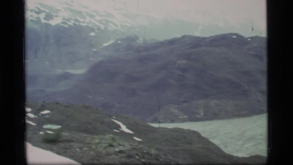 Soft 8mm frame of Glacier Bay with layered rocky slopes, pale blue fjord water, snow patches, and distant mountains under cloud.