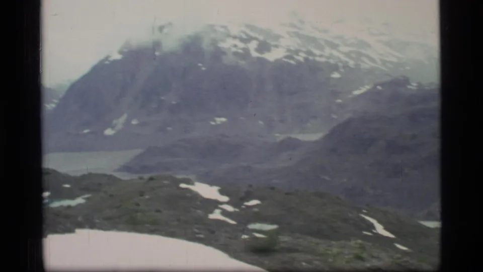 1977 Glacier Bay 8mm view of a snow-streaked mountain beyond rocky foreground slopes and glimpses of fjord water.