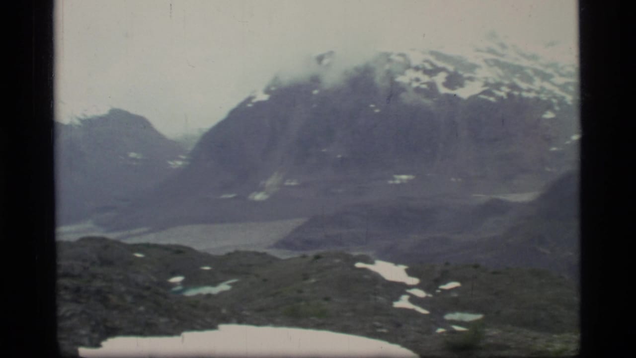 1977 8mm view of Glacier Bay mountains with snow patches, a rocky foreground, and pale water or ice in the valley below.