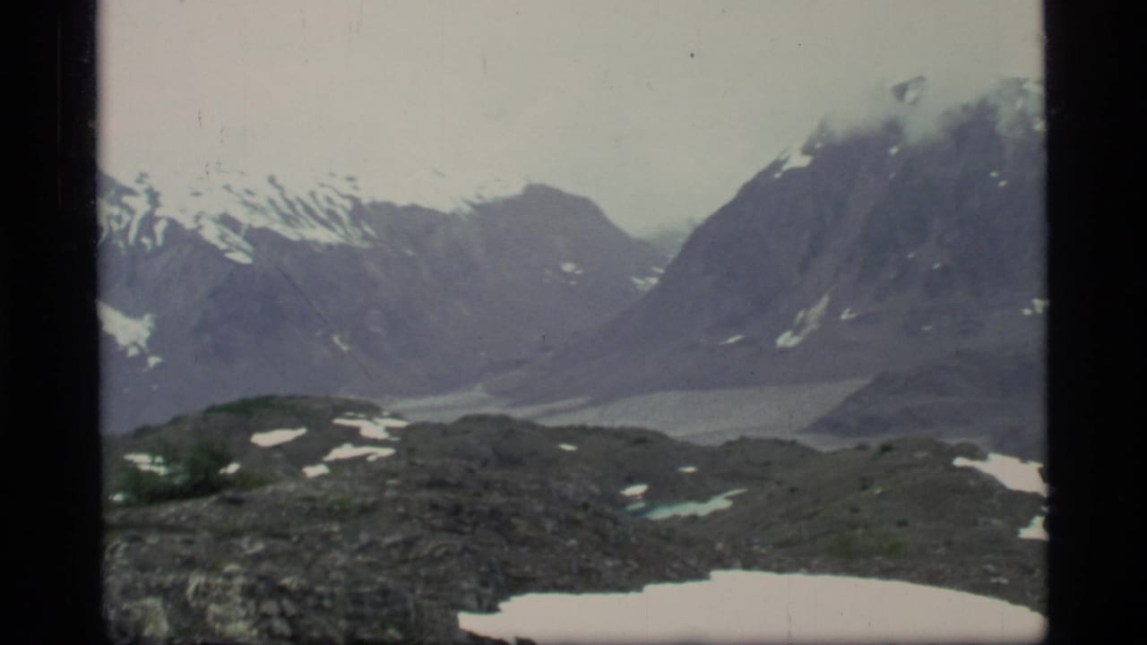A handheld 8mm Glacier Bay landscape view shows steep mountains, snow patches, and a pale glacial valley or lake below.